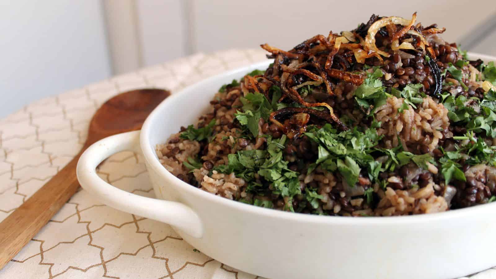 A white dish filled with rice, lentils, and caramelized onions is garnished with fresh cilantro. A wooden spoon lies beside the dish on a patterned fabric.