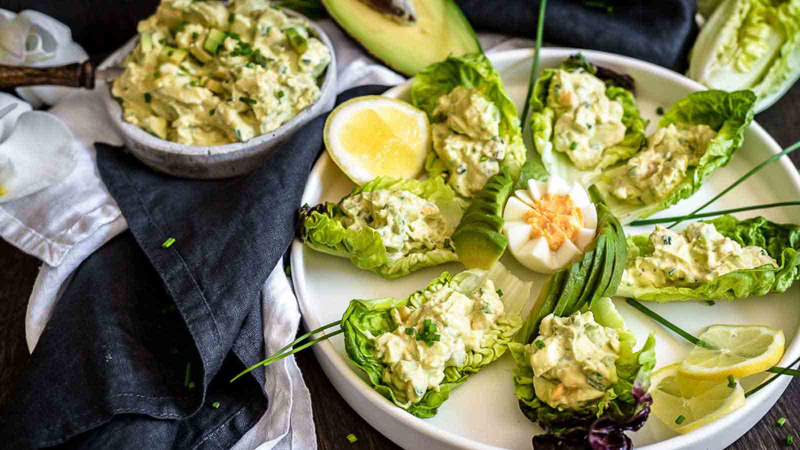 A plate with lettuce leaves filled with avocado egg salad, garnished with fresh herbs. A halved boiled egg and lemon slice rest on the side, resembling an auto draft from nature’s kitchen. In the background, a bowl with more salad joins an avocado half and a dark cloth napkin.