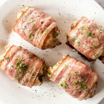 Four bacon-wrapped stuffed chicken breasts are placed on a white plate. The chicken is garnished with chopped herbs and pepper, set against a light marble background.