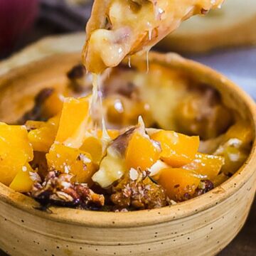 A wooden bowl filled with a dish featuring melted cheese, diced yellow peppers, and pecans. A chunk of cheese is being lifted from the bowl, showing its stretchy texture. Slices of bread and pecans are scattered around the bowl.
