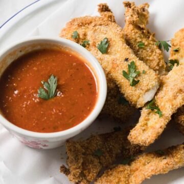 A plate with breaded fish sticks garnished with parsley, served alongside a small bowl of red dipping sauce, placed on a piece of white parchment paper.