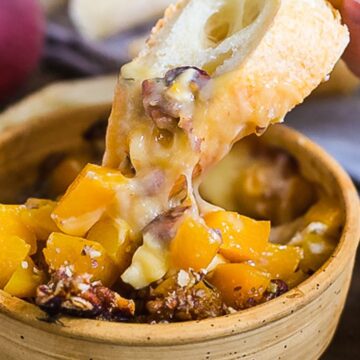 A close-up of a bowl containing melted cheese with chunks of peaches and pecans. A hand is dipping a slice of bread into the cheese mixture. Pieces of bread and pecans are scattered around the bowl on a wooden surface.