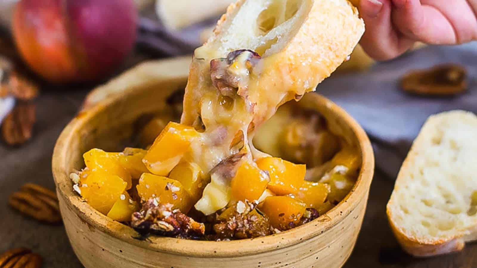 A person is dipping a piece of bread into a bowl of peach and pecan dip.