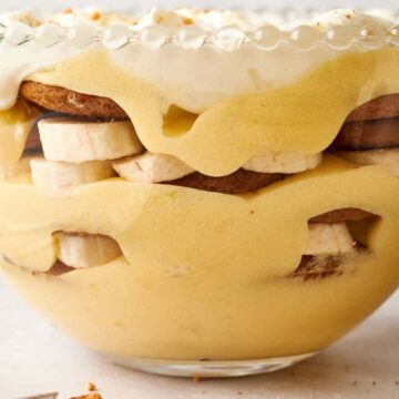 A glass bowl filled with layers of banana slices, cookies, and creamy custard topped with whipped cream. The layers are visible through the clear bowl, showcasing the dessert's textures and colors. Forks are seen to the side on the table.