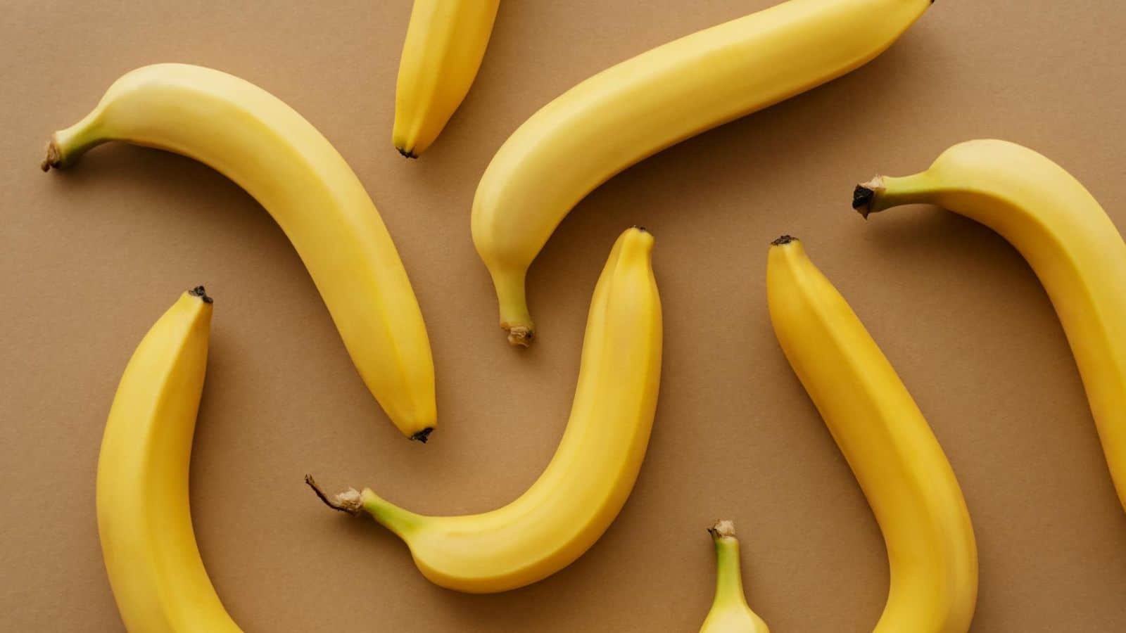 A flat lay image of seven yellow bananas arranged in a scattered pattern on a tan background. The bananas are evenly spaced and positioned at various angles, displaying their curved shapes.