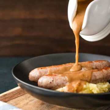 A black plate with two sausages on a bed of mashed potatoes. Brown gravy is being poured over the sausages from a white gravy boat. The background is dark with a wooden surface, and a blue and white checkered cloth is partially visible to the side.