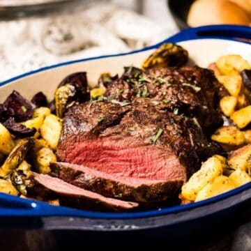 A blue roasting dish contains a cooked beef roast surrounded by roasted potatoes and purple vegetables. The beef is sliced to show a pink interior. The dish is placed on a wooden surface, and bread rolls are visible in the background.