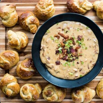 A black bowl of creamy dip topped with bacon bits and green onions sits on a wooden board, surrounded by golden-brown pretzel knots. A silver spoon and a folded gray napkin are placed beside the board.