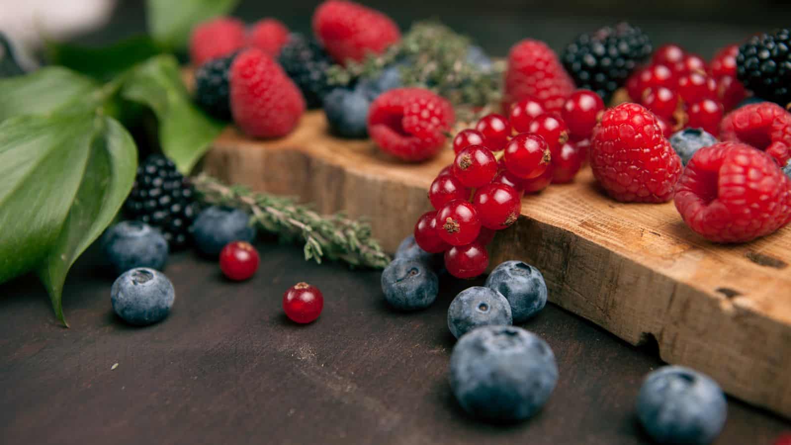 A wooden board with an assortment of berries, including raspberries, red currants, blackberries, and blueberries. Some thyme and green leaves are also visible on the dark surface.