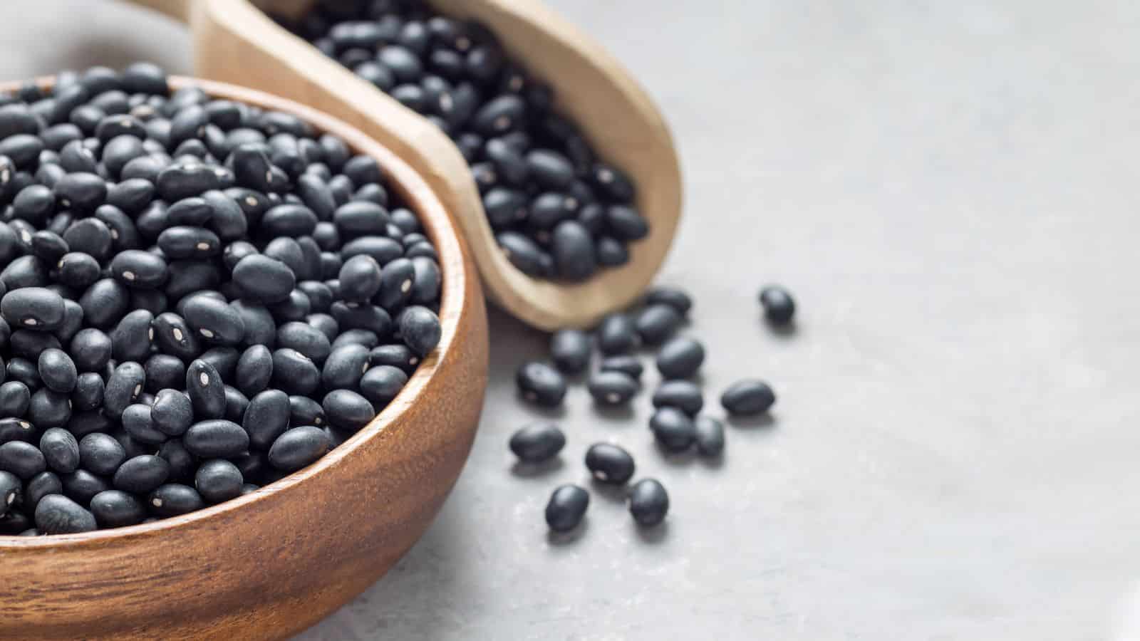 A wooden bowl filled with black beans is placed on a gray surface. A wooden scoop lies beside it, spilling a few beans onto the surface. The beans are small, shiny, and round.