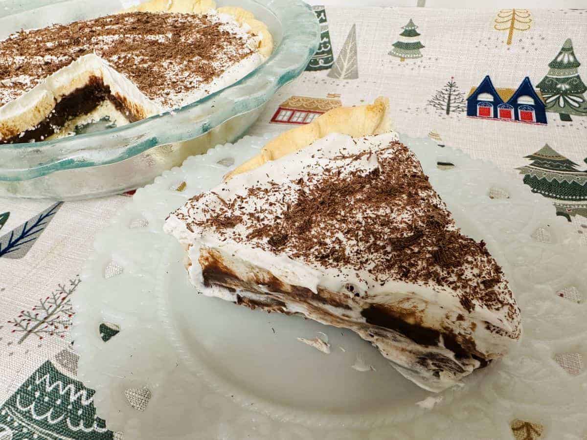 A slice of chocolate cream pie with a whipped topping and chocolate shavings sits on a decorative plate, perfect for those seeking easy pies. In the background, a holiday-themed tablecloth features charming illustrations, while another pie is partially visible in a glass dish.