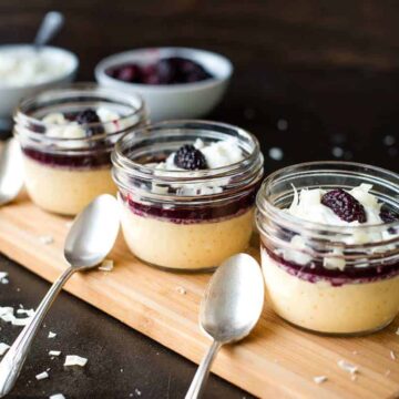 Three small glass jars of yellow pudding, crowned with dark berry compote and white chocolate shavings, rest on a wooden board—perfect festive desserts. Three silver spoons sit in front, with a bowl of cream and berries creating a tempting backdrop.