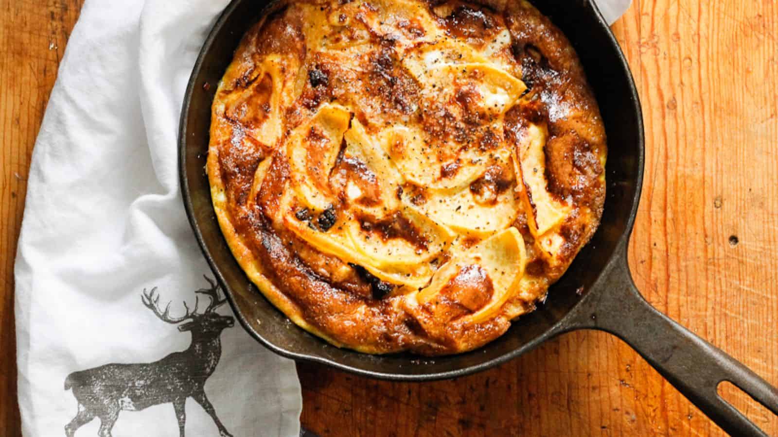 A baked dish, possibly a frittata or omelet, with slices of fruit on top, sits in a black cast iron skillet on a wooden surface. A white cloth with a black deer illustration is partially visible beside it.