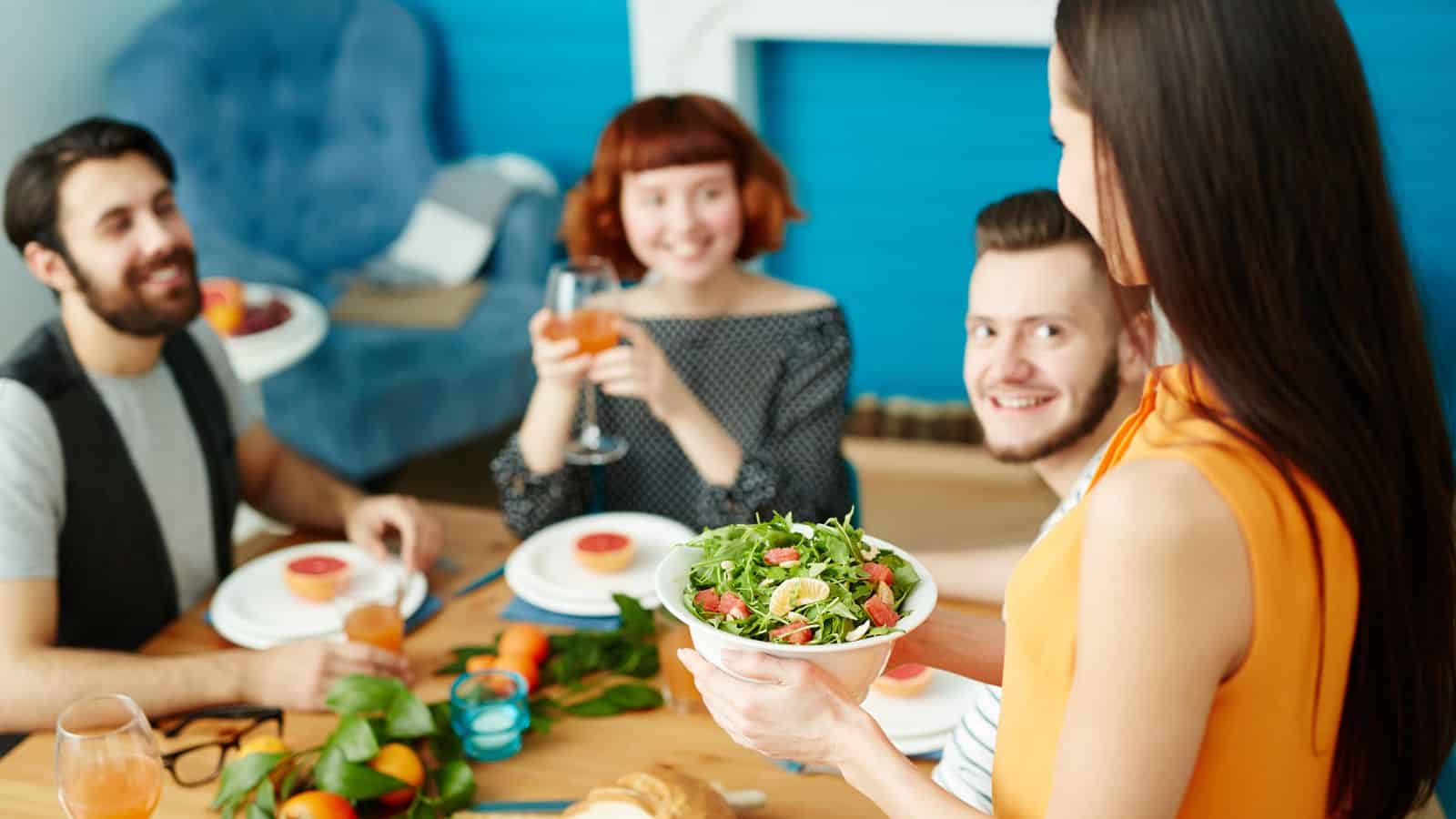 A group of four people sits around a dining table with colorful plates and drinks, enjoying a healthier holiday meal. A woman in the foreground serves a bowl of salad with simple tips for wellness in mind. The scene is set in a bright, cozy room with blue accents on the walls and chairs.