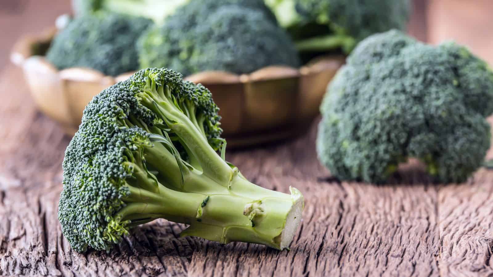 Fresh broccoli florets are placed on a rustic wooden surface. A small bowl in the background contains more broccoli, and the focus is on the detailed texture of the broccoli in the foreground.