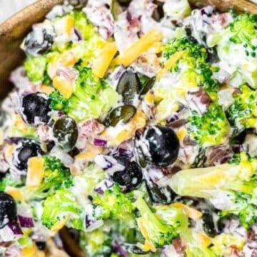 A wooden bowl filled with a broccoli salad that has everyone asking, "Did you make this?" It features chopped broccoli, black olives, shredded cheese, sunflower seeds, diced red onion, and a creamy dressing—perfect for holiday salads.