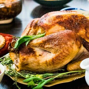 A roasted turkey garnished with herbs is placed on a platter. Surrounding the turkey are various dishes, including cranberry sauce, a pie, and a bowl of green beans. A white gravy boat is also visible in the foreground.