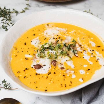 A white bowl filled with creamy pumpkin soup garnished with a drizzle of cream, chopped nuts, and fresh thyme. The bowl is placed on a marble surface next to a small wooden bowl of nuts and sprigs of thyme. A gray cloth lies nearby.