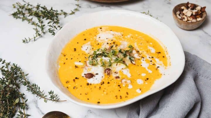A white bowl filled with creamy pumpkin soup garnished with a drizzle of cream, chopped nuts, and fresh thyme. The bowl is placed on a marble surface next to a small wooden bowl of nuts and sprigs of thyme. A gray cloth lies nearby.