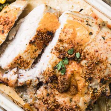 Sliced roasted turkey breast in a baking dish, seasoned with herbs and spices. Surrounded by cooked green beans, corn, and onions. Garnished with fresh herbs.