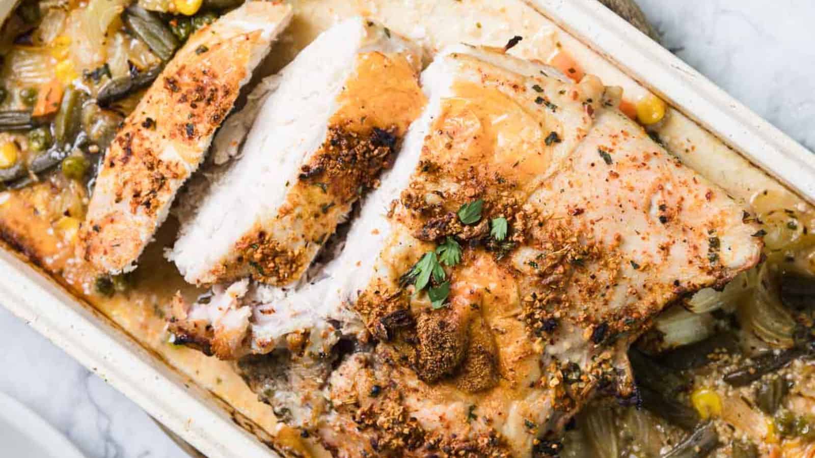 Sliced roasted turkey breast in a baking dish, seasoned with herbs and spices. Surrounded by cooked green beans, corn, and onions. Garnished with fresh herbs.