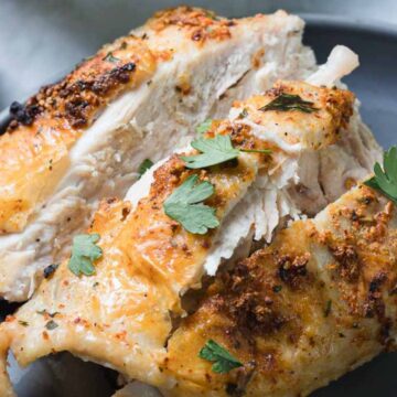 A plate with pieces of roasted chicken, seasoned with herbs and spices, garnished with fresh parsley leaves. A checkered cloth is visible in the background.