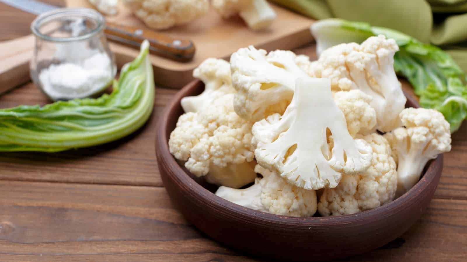 A brown bowl filled with cauliflower florets is placed on a wooden table. A small jar of salt and a knife on a cutting board with additional cauliflower are in the background. Green leaves are scattered around.