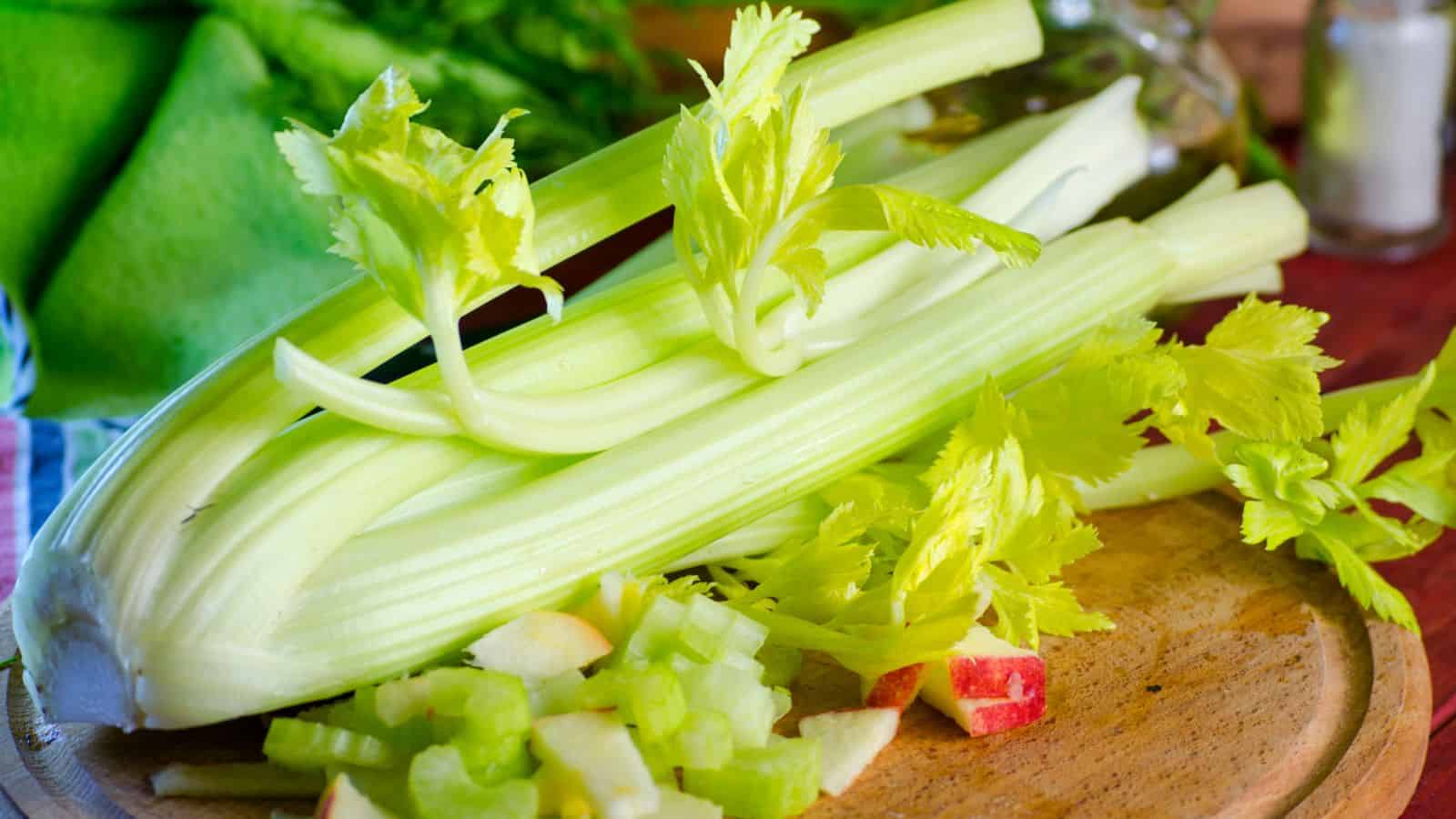 A bunch of fresh celery with leaves is placed on a wooden cutting board. Several celery stalks are chopped next to apple chunks. A leafy green is in the background along with a glass bottle and a salt shaker.