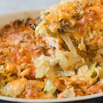 A close-up of a baked cheesy dish with shredded cheese and bread crumbs on top. A serving is being lifted, showing melted cheese stretching above the baking dish. The dish has a crispy, golden-brown crust.