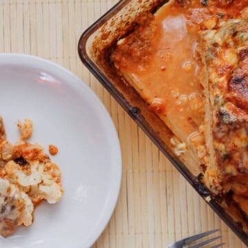 A baked cauliflower dish with melted cheese and tomato sauce is on a rectangular glass baking dish. A portion is served on a white plate beside it. A fork rests on the table near the plate.