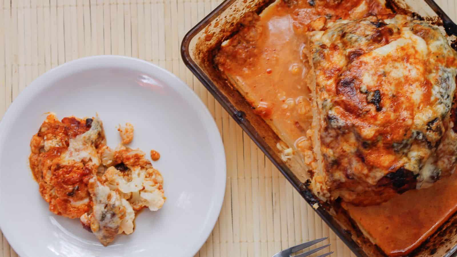 A baked cauliflower dish with melted cheese and tomato sauce is on a rectangular glass baking dish. A portion is served on a white plate beside it. A fork rests on the table near the plate.