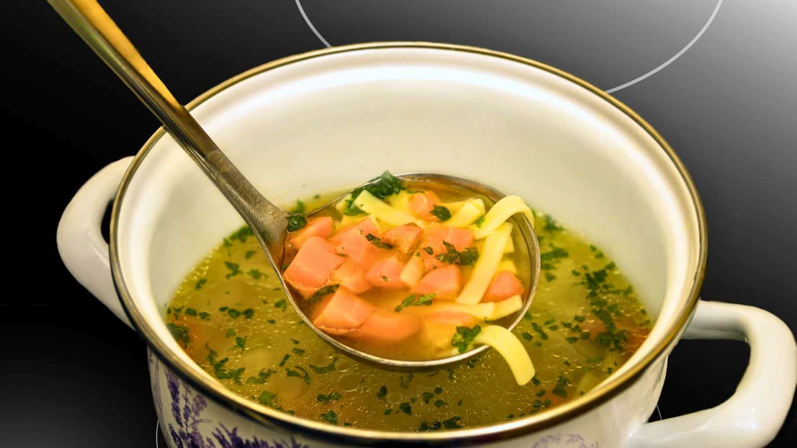 A ladle holds a portion of soup above a pot. The soup contains diced pink sausages, yellow noodles, and green herbs in a clear broth. The pot is white with a floral design and has handles. The background is dark.
