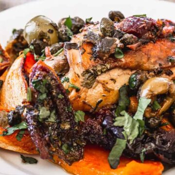 A plate of roasted chicken topped with capers and herbs, surrounded by roasted vegetables including green olives, sliced carrots, and wedges of sweet potatoes. A fork rests to the side on the white plate.