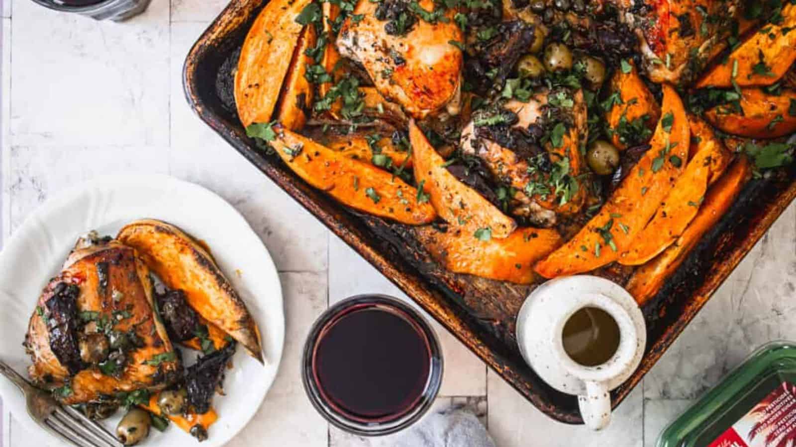 A roasted chicken and sweet potato dish garnished with herbs sits on a baking tray. Beside it, a plate offers a serving of the same meal. A glass of red wine, a white mug, and a cloth napkin are nearby on a light-colored tabletop.