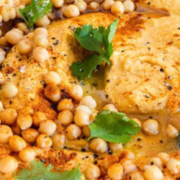 A close-up of a plate of hummus topped with chickpeas, olive oil, paprika, black pepper, and garnished with fresh cilantro leaves.