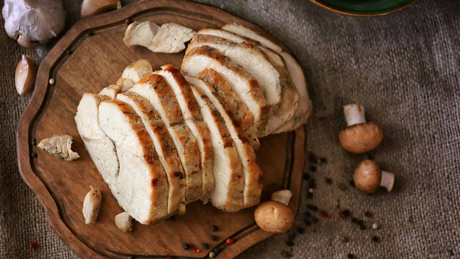 Sliced roasted pork on a wooden platter, garnished with garlic cloves, peppercorns, and small mushrooms, captures the essence of a holiday feast. This sumptuous spread is artfully arranged on a textured fabric surface to enhance the festive dining experience.