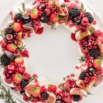 A holiday wreath-shaped dessert topped with a variety of fresh berries, figs, and pomegranate seeds arranged on a white plate. Sprigs of rosemary add a decorative touch. Plates with more berries and pomegranate seeds are nearby.