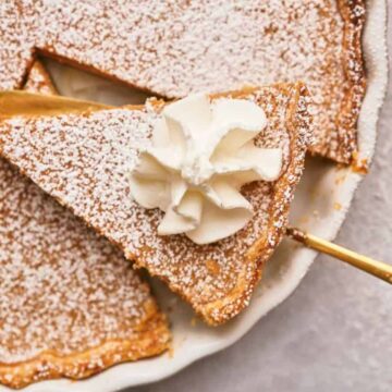 A close-up of a sliced pie on a dish, with a piece being lifted by a golden serving utensil. The pie is topped with powdered sugar and a dollop of whipped cream on one slice.