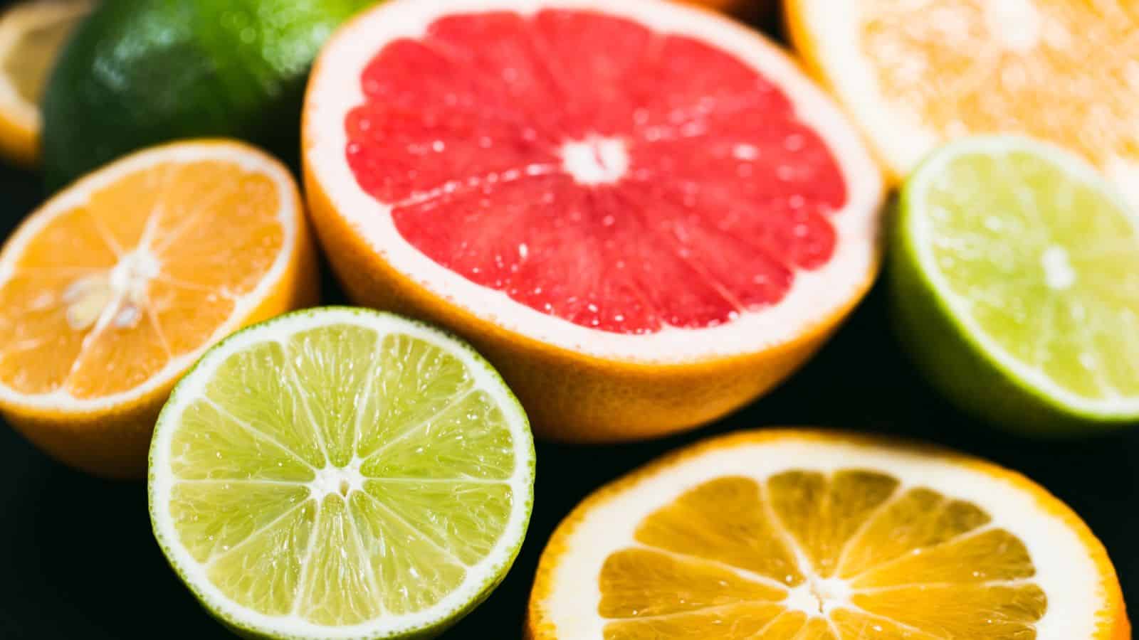 Close-up image of various halved citrus fruits, including a red grapefruit, orange, lemon, and lime. The fruits are arranged closely together on a dark surface, showcasing their vibrant colors and juicy textures.