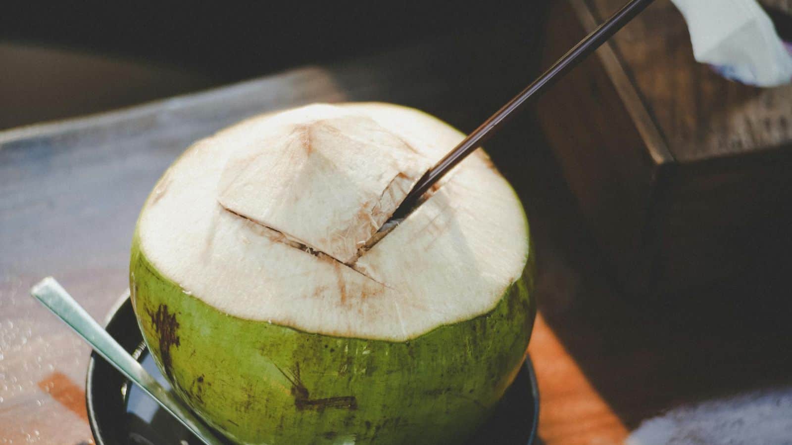 A fresh green coconut with the top cut open, featuring a straw for drinking. It is placed on a plate with sunlight casting shadows on the wooden surface beneath.