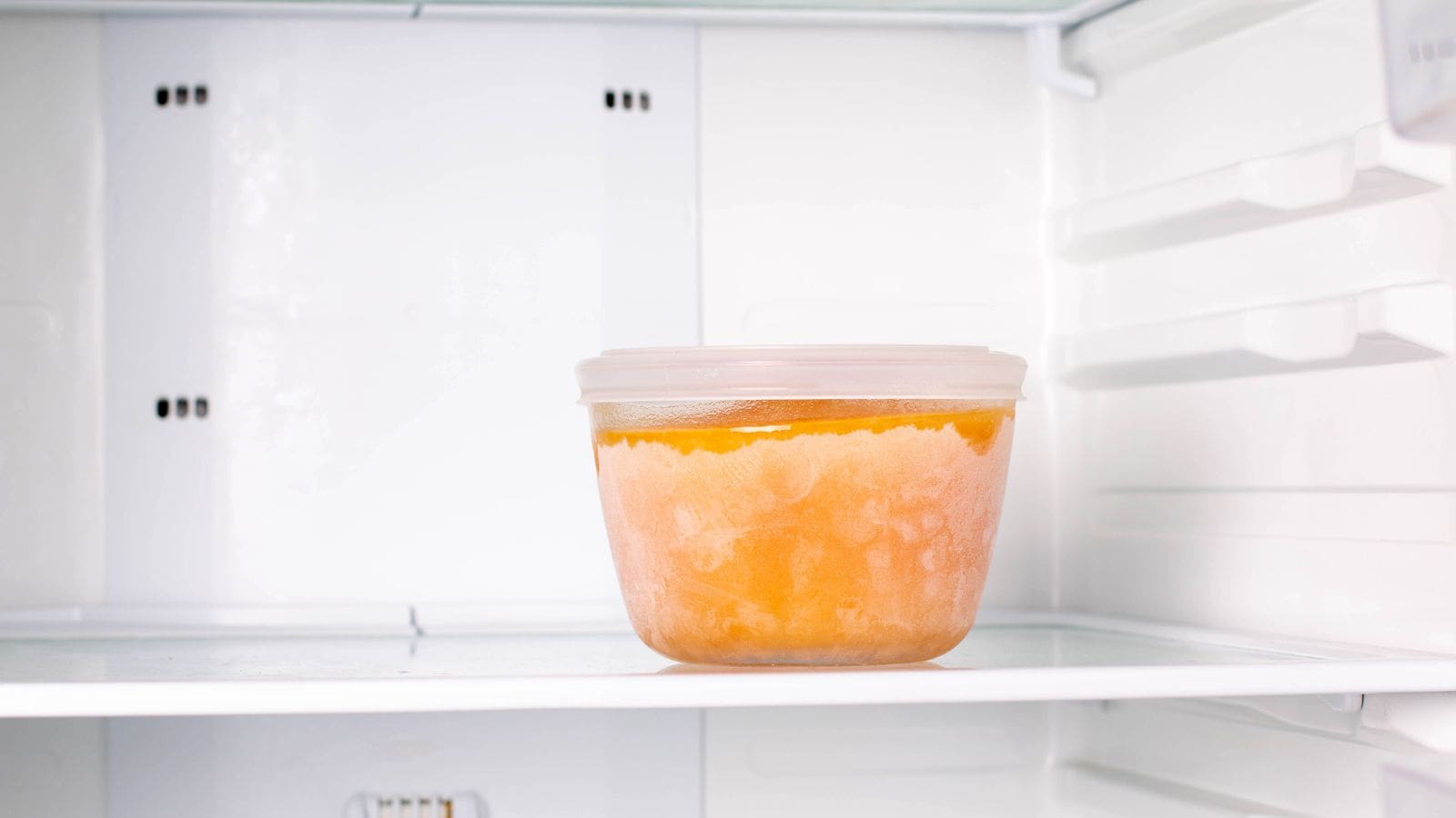 A clear plastic container filled with an orange, frozen substance sits on the middle shelf of an otherwise empty white refrigerator. Perfectly suited for holiday meal planning, this neatly labeled container with a white lid highlights the clean and well-lit fridge interior.