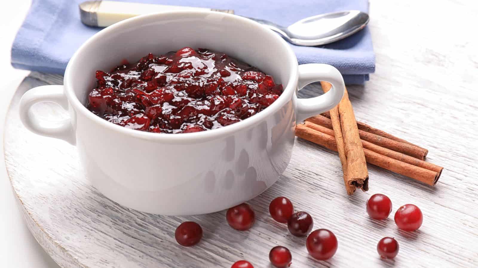 A white bowl filled with cranberry sauce is placed on a light wooden surface. Around the bowl are scattered fresh cranberries and two cinnamon sticks. A spoon and a folded blue napkin are positioned nearby.