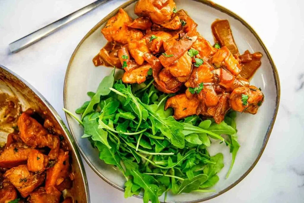 A white plate featuring chicken curry garnished with herbs sits alongside a bed of fresh arugula. Nearby, part of the dish is visible in a pan, ready to serve. A silver fork rests on the table, completing this effortlessly curated auto draft culinary masterpiece.