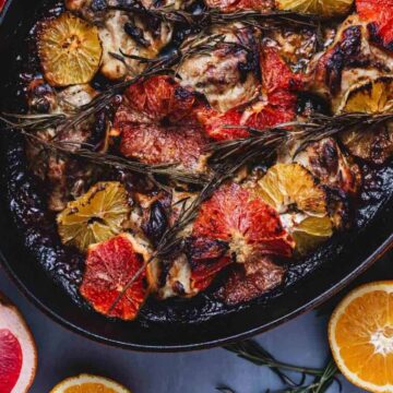 A roasted dish in a pan featuring various cuts of meat topped with citrus slices and rosemary sprigs. Surrounding the pan are halved grapefruits, lemons, and oranges on a blue surface.