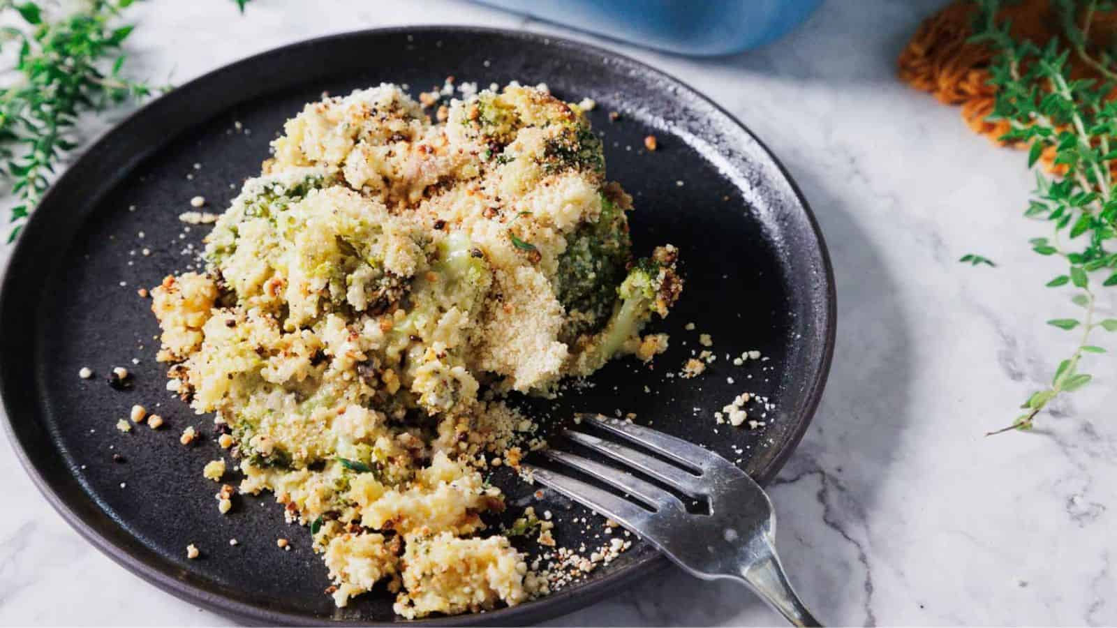 A black plate holds a serving of baked broccoli casserole topped with breadcrumbs. A fork lies beside the food on the plate. The background has a marble surface with greenery partially visible.