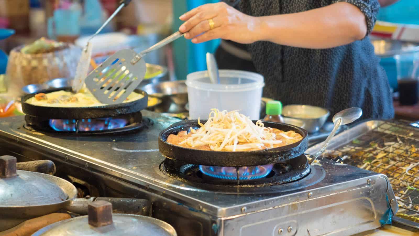 In the midst of holiday meal planning, a person cooks food on a stovetop, skillfully using a spatula to prepare dishes in round pans. One pan is filled with noodles, while another holds a different culinary delight. The kitchen bustles with utensils and ingredients hinting at festive creativity.