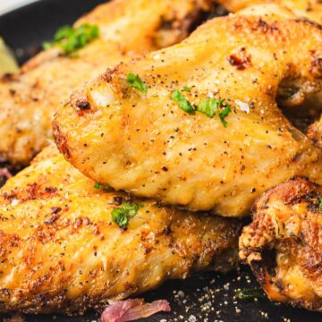 A plate of seasoned and grilled chicken wings garnished with chopped herbs. Lemon wedges are visible in the background. The wings have a crispy, golden-brown texture.