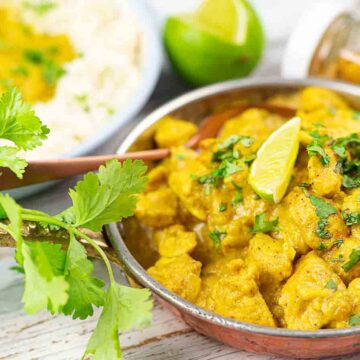 A bowl of chicken curry with tender meat chunks, garnished with cilantro and a lime wedge, is a crowd-pleasing dish perfect for family gatherings. A spoon rests in the curry, while extra curry and fluffy white rice await in the background, accompanied by fresh cilantro and a small jar of spices on the light wooden surface.