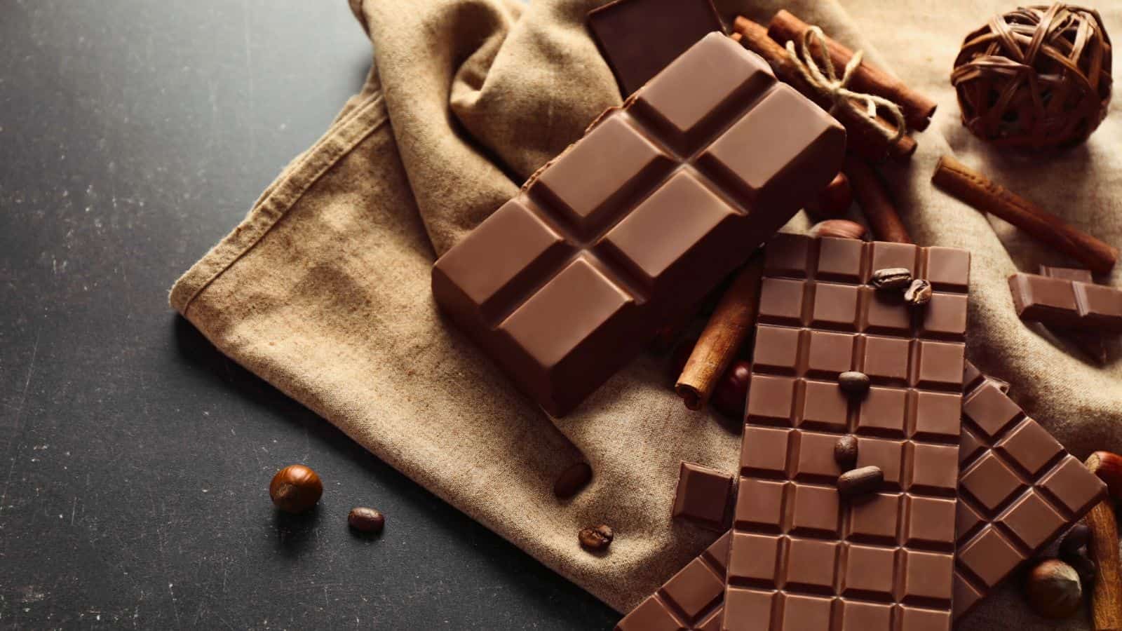 Chocolate bars and pieces are arranged on a fabric cloth, surrounded by coffee beans, nuts, and cinnamon sticks on a dark surface. A decorative wicker ball is also visible.