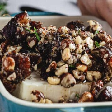 Hands holding a dish with a block of cheese topped with a mixture of nuts, herbs, and dates. The ingredients are layered in a blue rectangular dish.